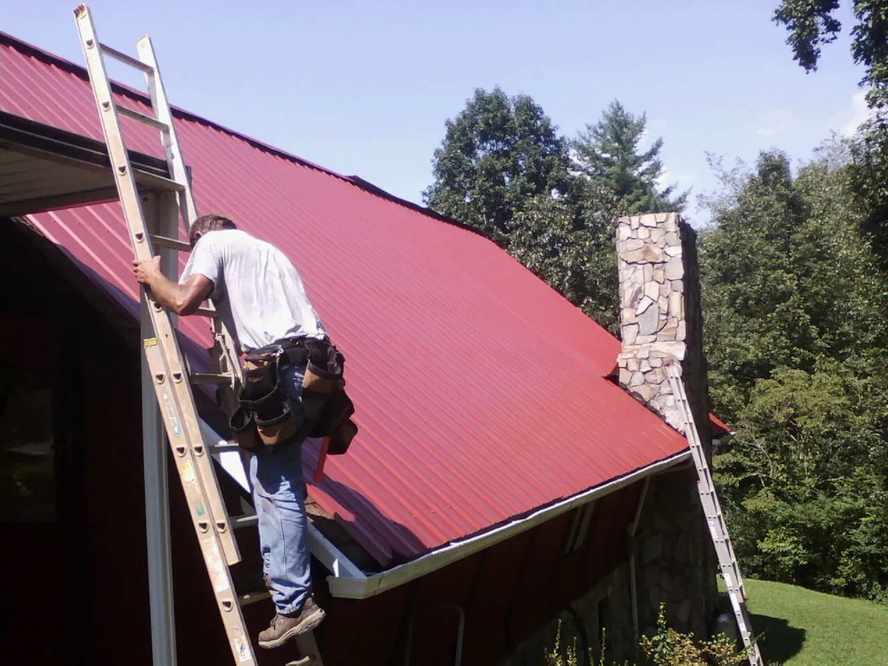 Roofer on a ladder beside a red metal roof and stone chimney — Soffit & Fascia Repair by Roofing Brothers in Winona